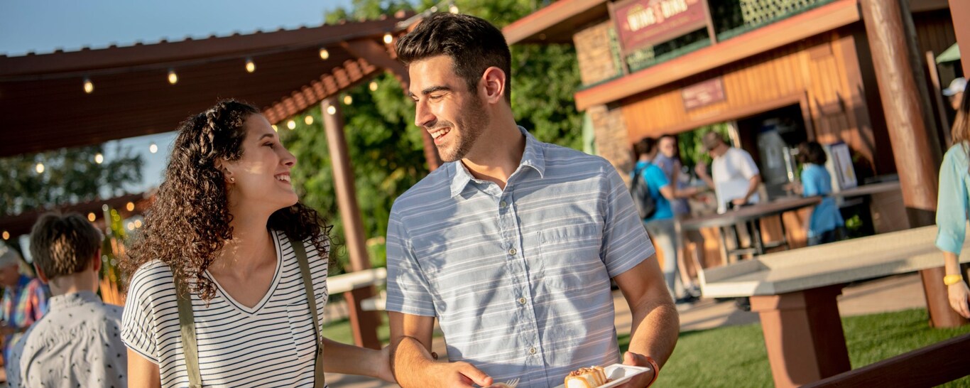 Un hombre y una mujer sostienen pequeños platos de comida y sonríen mientras pasan por un puesto de los Mercados del Mundo