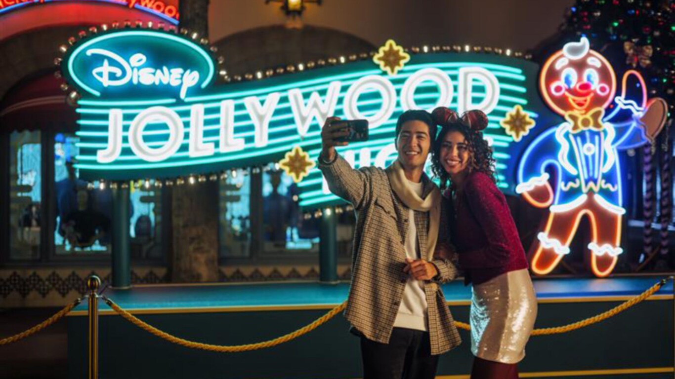 A couple takes a selfie in front of a neon sign that reads ‘Disney Jollywood Nights’