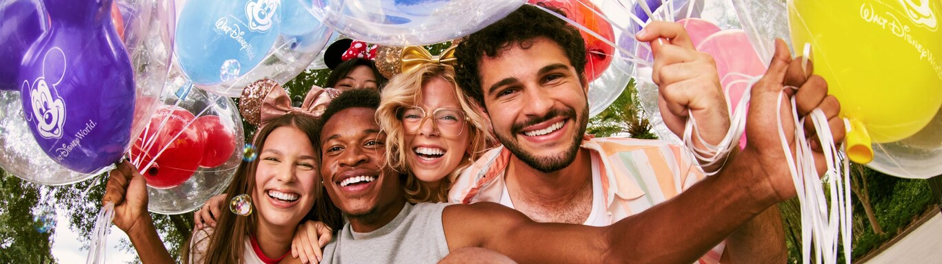 A group of five friends sit closely together, smiling joyfully, surrounded by Mickey Mouse shaped balloons