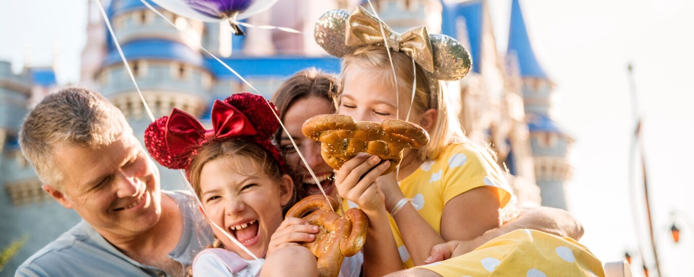 Una familia sonríe y se abraza mientras sostiene globos y pretzels de Mickey Mouse frente al Cinderella Castle