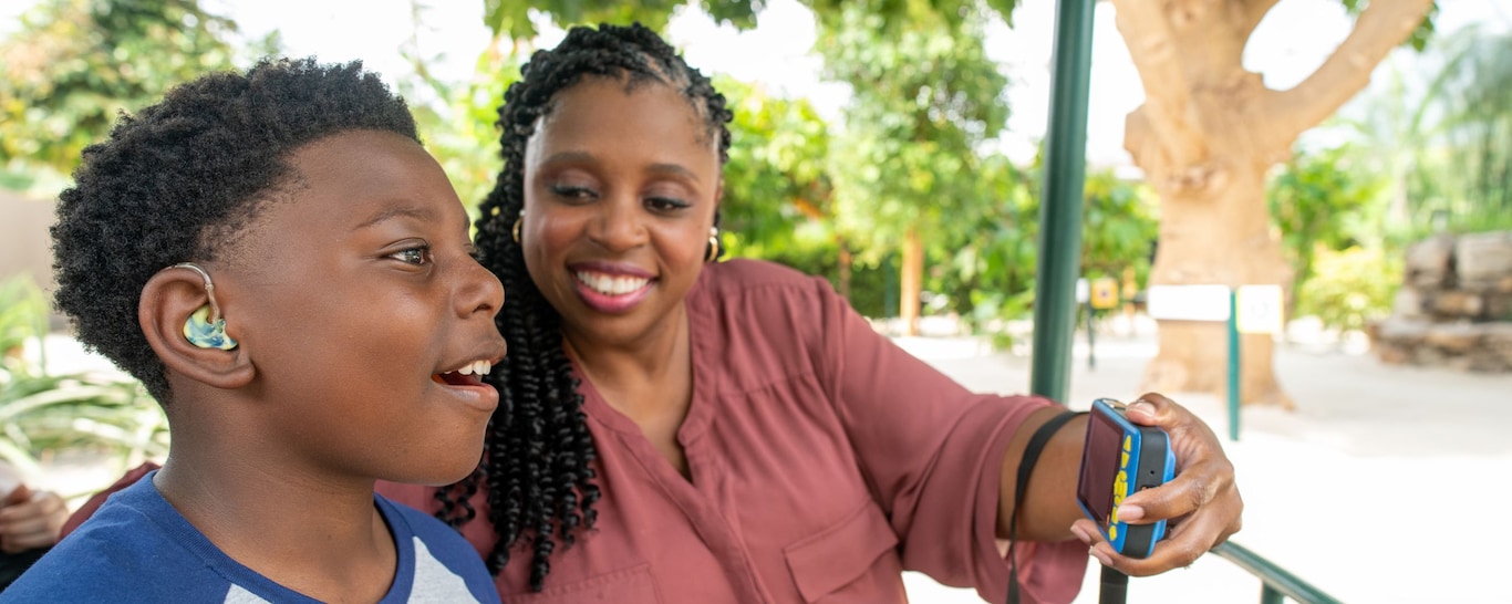A boy with a hearing aid looks in awe while a woman holds the Disney Handheld Device in front of him.
