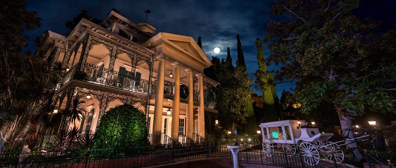 At nighttime, a horse drawn carriage sits outside the eerily illuminated exterior of the Haunted Mansion at Disneyland Park theme park in California