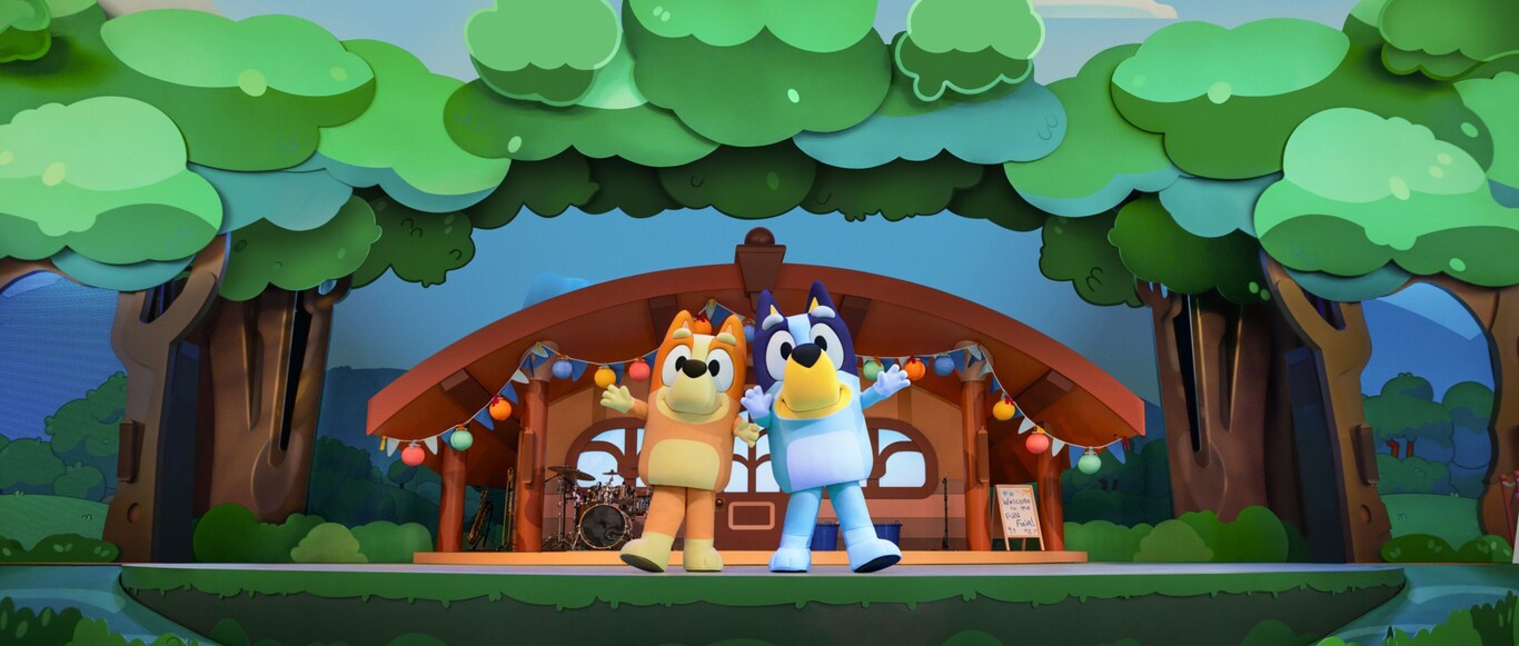 Bingo and Bluey pose on a stage set designed to look like a house in the woods at Bluey’s Best Day Ever