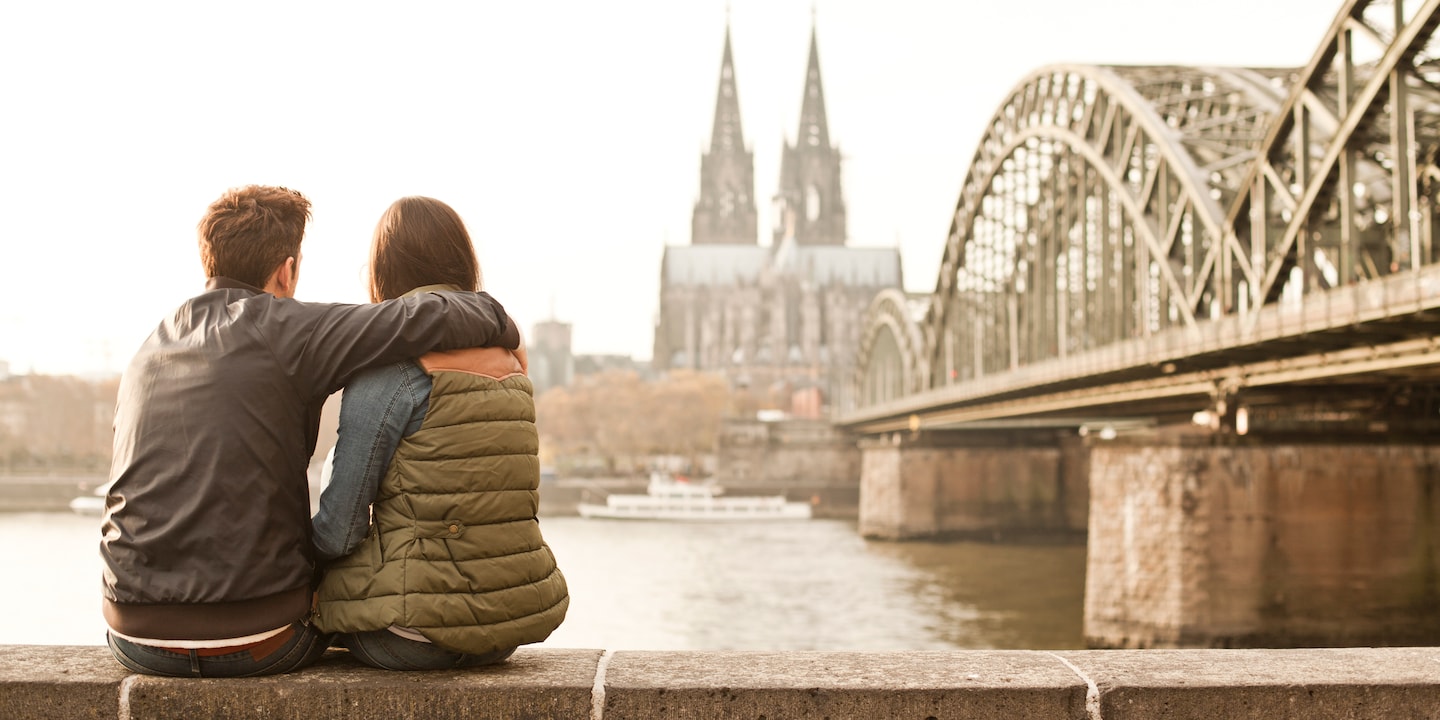 A couple sit on a bench looking out over the Rhine with the Cologne Cathedral in the background