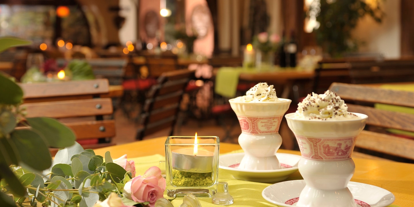 Two cups, topped with whipped cream, sit on a table with a lit candle and several roses in a restaurant with wood slatted chairs