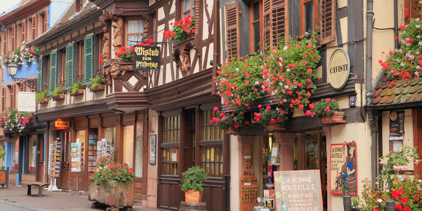 Several storefronts in traditional French style, with flowers in window boxes, line a quaint street in Riquewihr, France