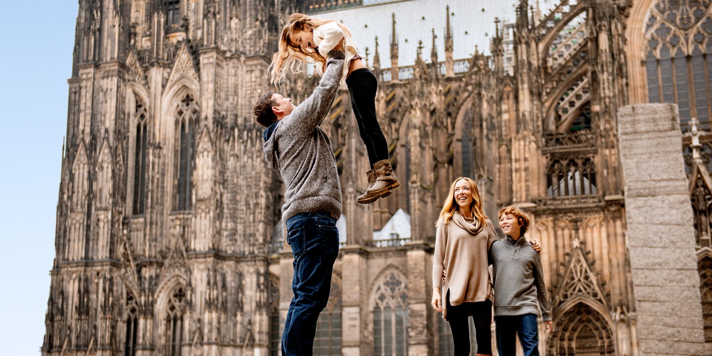 A father lifts his daughter up high in front of the Cologne Cathedral as his wife and son watch