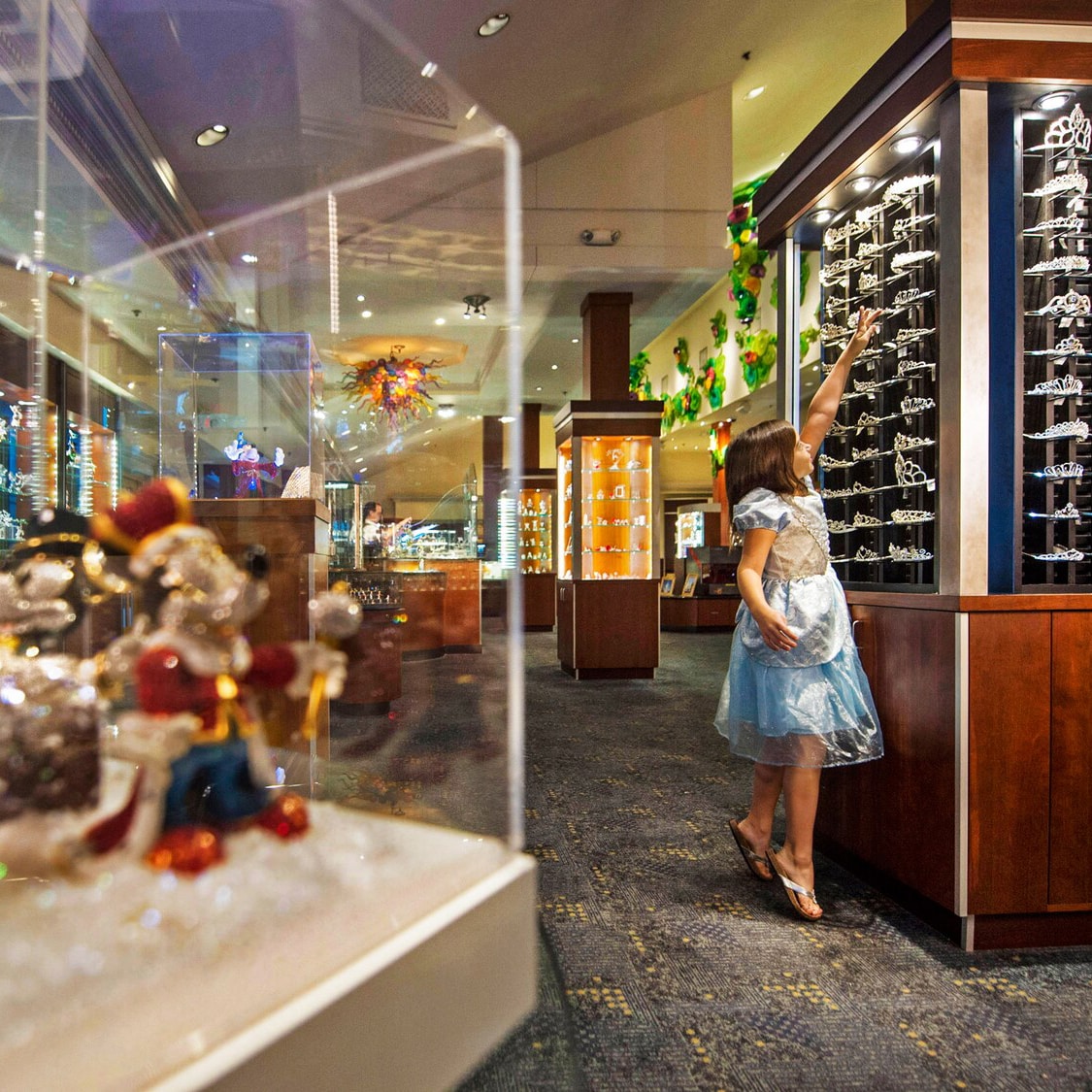 A young girl in a princess dress stands on her tiptoes to reach a tiara in a display case