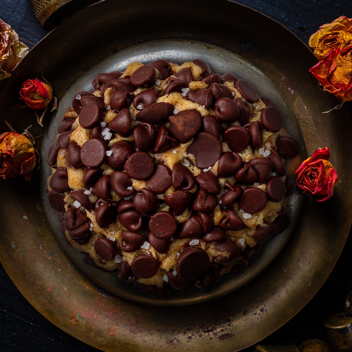 A chocolate chip cookie rests on a brass plate surrounded by dried roses