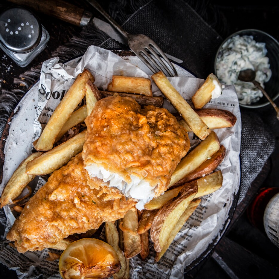 A plate of fish and chips rests on a table alongside utensils, a beverage, a salt shaker and a small bowl of tartar sauce