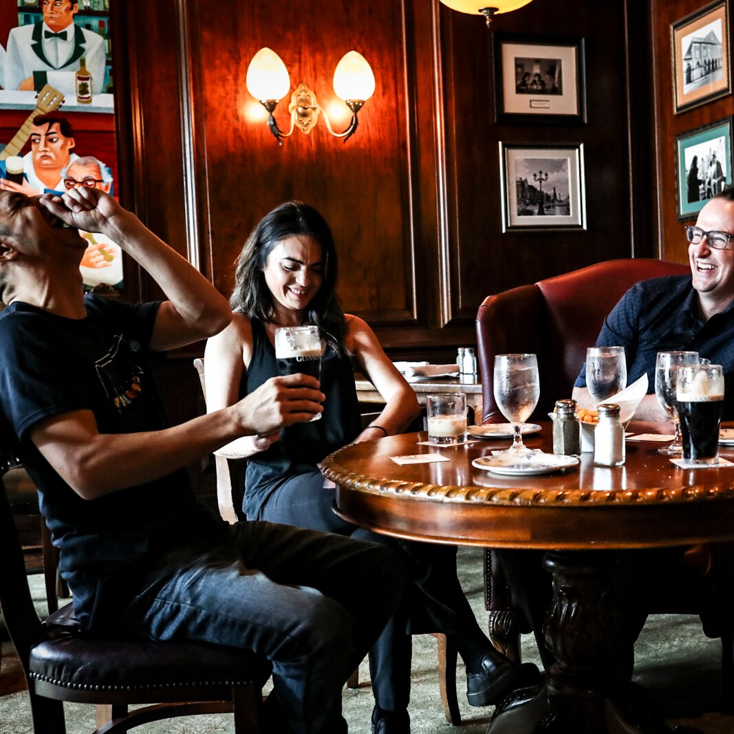 In a warmly decorated paneled room, 3 men and a woman are seated at a wood table with drinks and a basket of chips