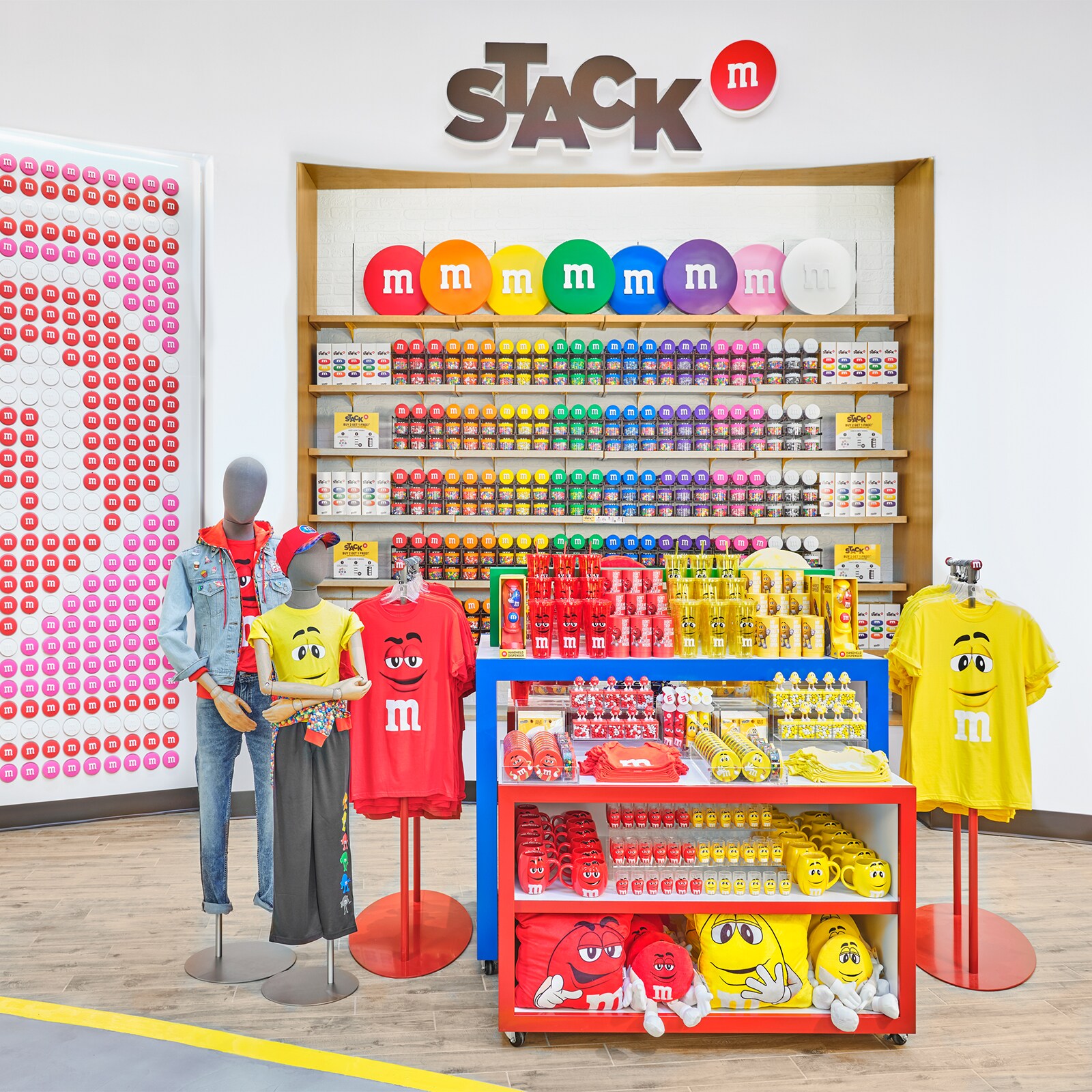 A large mosaic made up of tiles that resemble M and M candies, and display racks with M and M merchandise, including shirts, jackets, mugs, travel cups, pillows and packages of candy