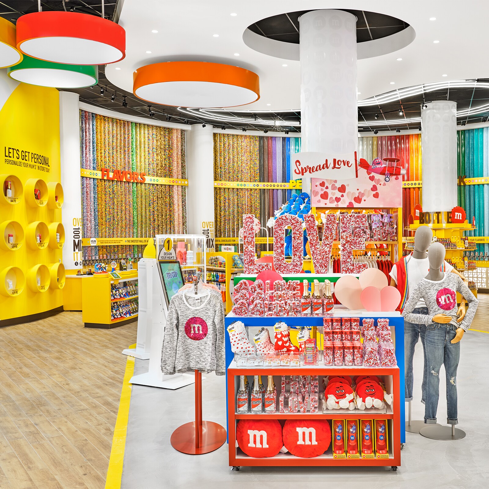 A store filled with M and M merchandise, including socks, shirts, toys, pillows, tall cylinders of candy and containers shaped like hearts, letters and Champaign bottles that are filled with candy. A sign shaped like the word Love is filled with candy. A touch screen kiosk has a sign that reads Design Your M and Ms.