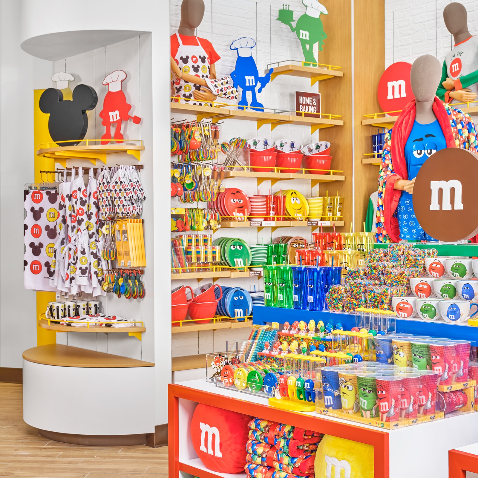 Display racks with M and M merchandise that is used in cooking, including bowls, measuring cups, plates, bowls, travel cups, whisks, oven mitts, cookie molds, straws and aprons. A sign reads Home and Baking.