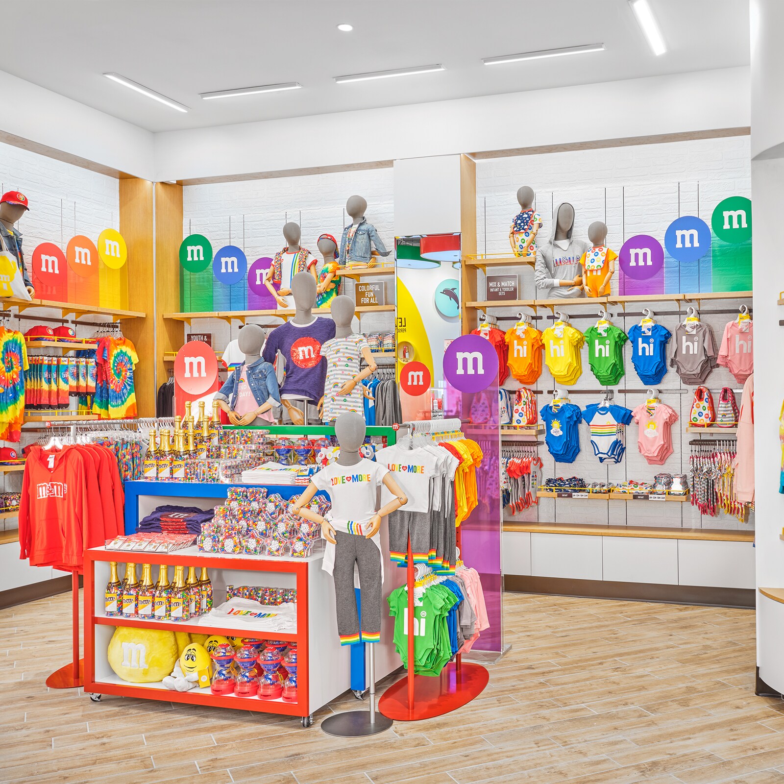Display racks with M and M merchandise for children, like shirts, pants, bibs, onesies, hats, toys, pillows and various containers filled with candy, including Champaign bottles