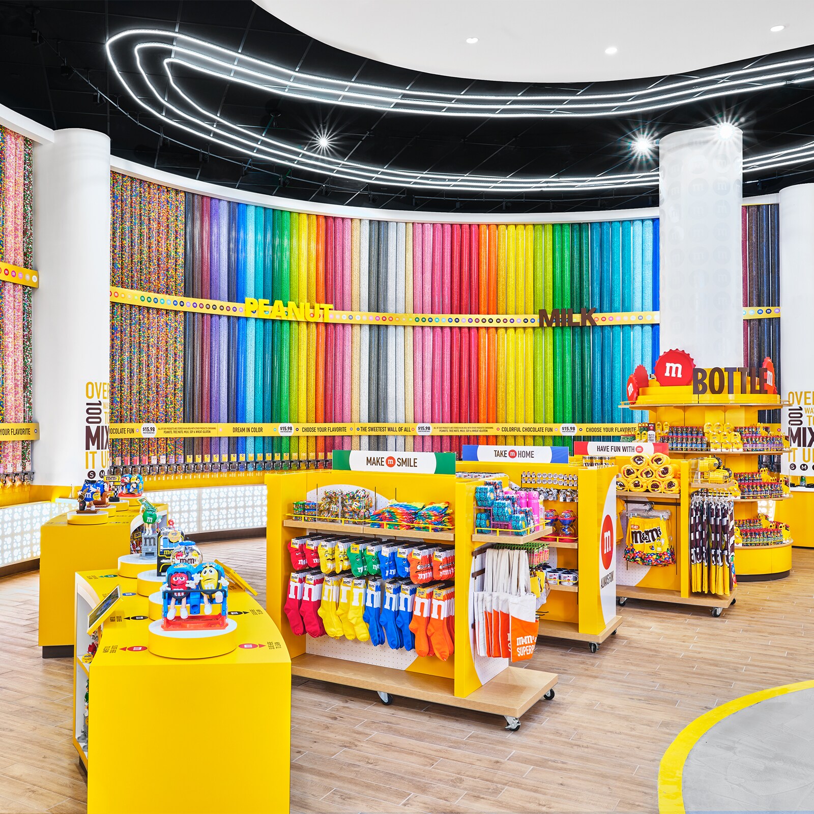 Tall cylinders of candy line the back wall of a store filled with M and M merchandise, including toys, socks, tote bags and towels. Signs read Flavors, Peanut, Milk, Make M Smile, Take M Home, Have Fun with M, and Bottle.