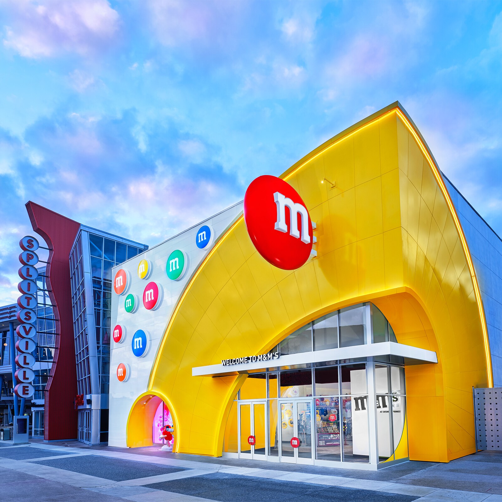 The exterior of a store with a marquee that reads Welcome to M and Ms, decorated with giant M and M candies, next door to a building with a sign that reads Splitsville