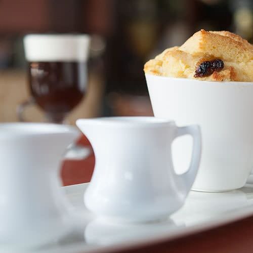 Bread and butter pudding in a ramekin next to 2 small jugs of sauce