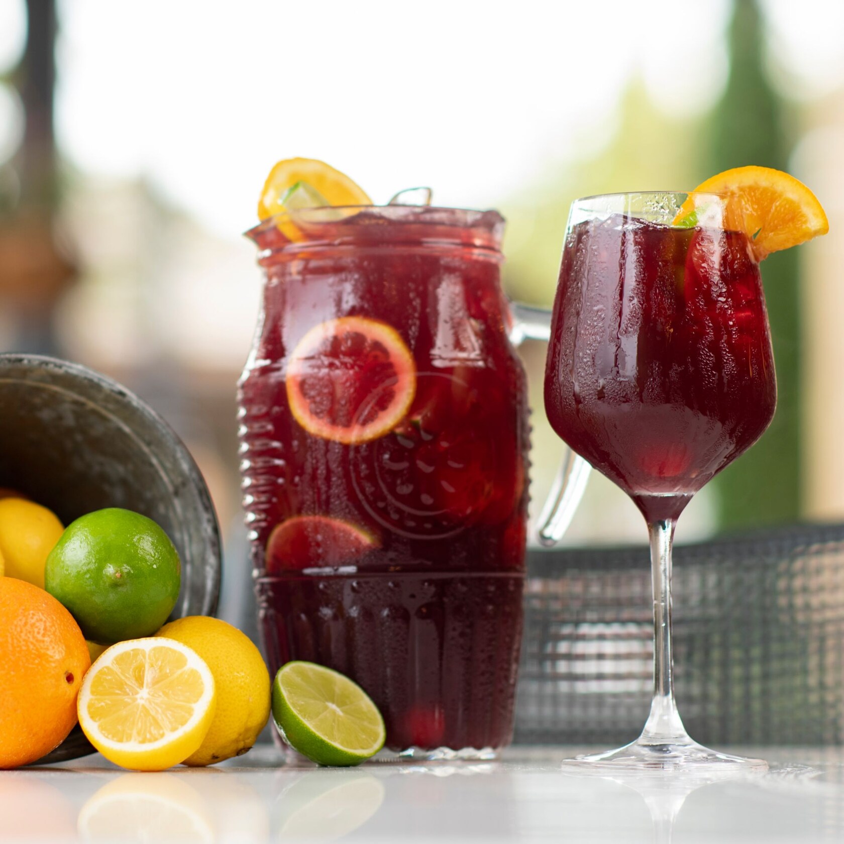 A glass of juice next to another container of juice and a bucket of lemons, oranges and limes