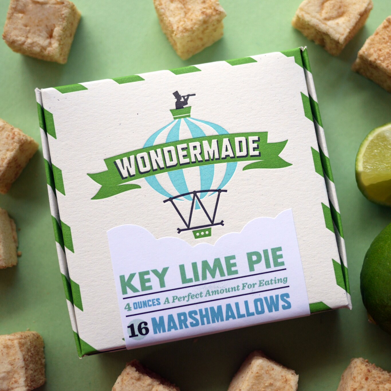Una caja de 16 malvaviscos Wondermade sabor Pastel de Limón Verde, rodeada de limón verde en rodajas y cuadrados de malvavisco cubiertos con migajas de galletas Graham.
