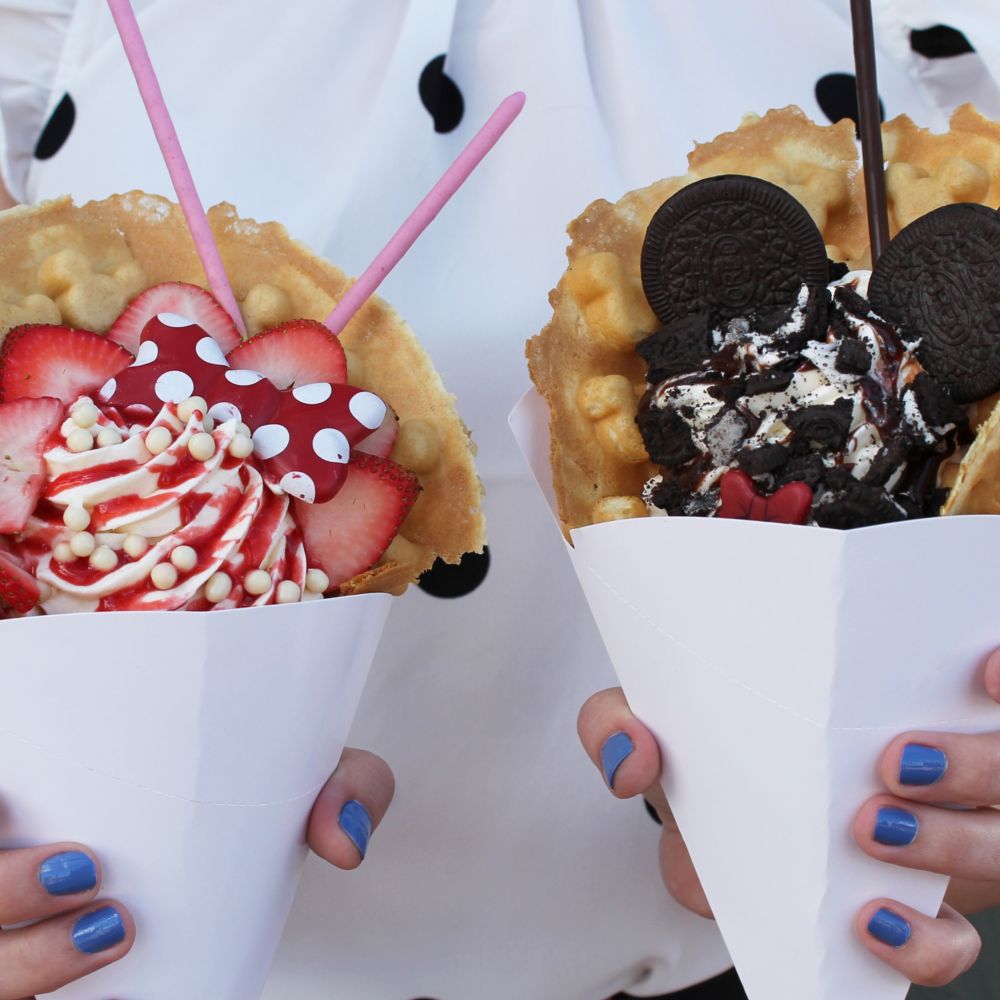 2 hands holding up waffle cones with ice cream and toppings 	