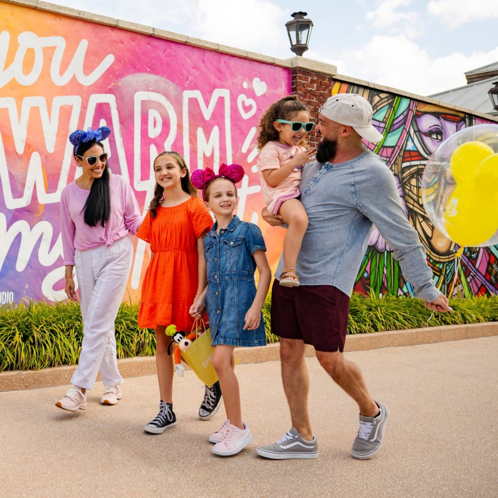 Una familia de 5 integrantes con un globo con forma de Mickey Mouse caminando frente a diferentes murales exhibidos en grandes paredes de ladrillo