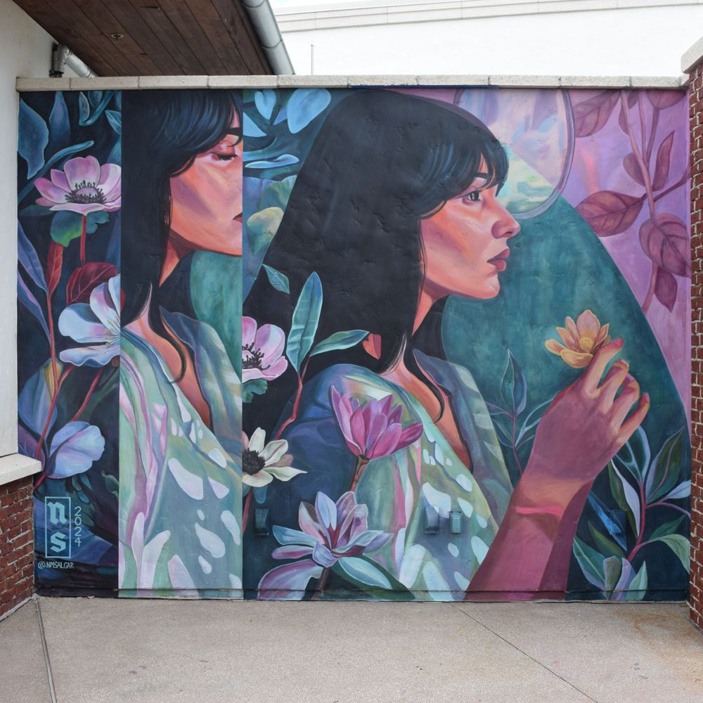A mural against a brick wall by Nicole Salgar featuring 2 profiles of a woman surrounded by flowers and foliage, holding a flower