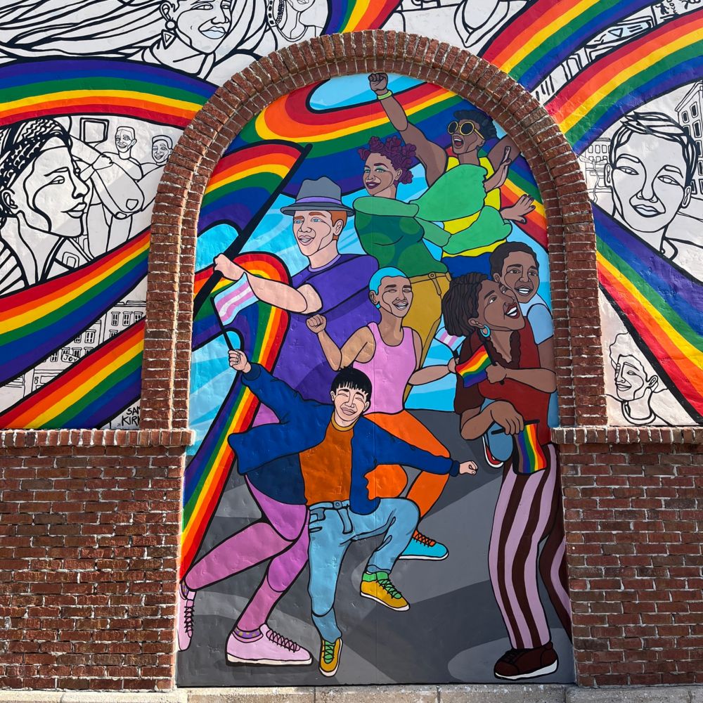 A mural by artist Sam Kirk featuring diverse people dancing under rainbows framed by brick arches and sketches of faces