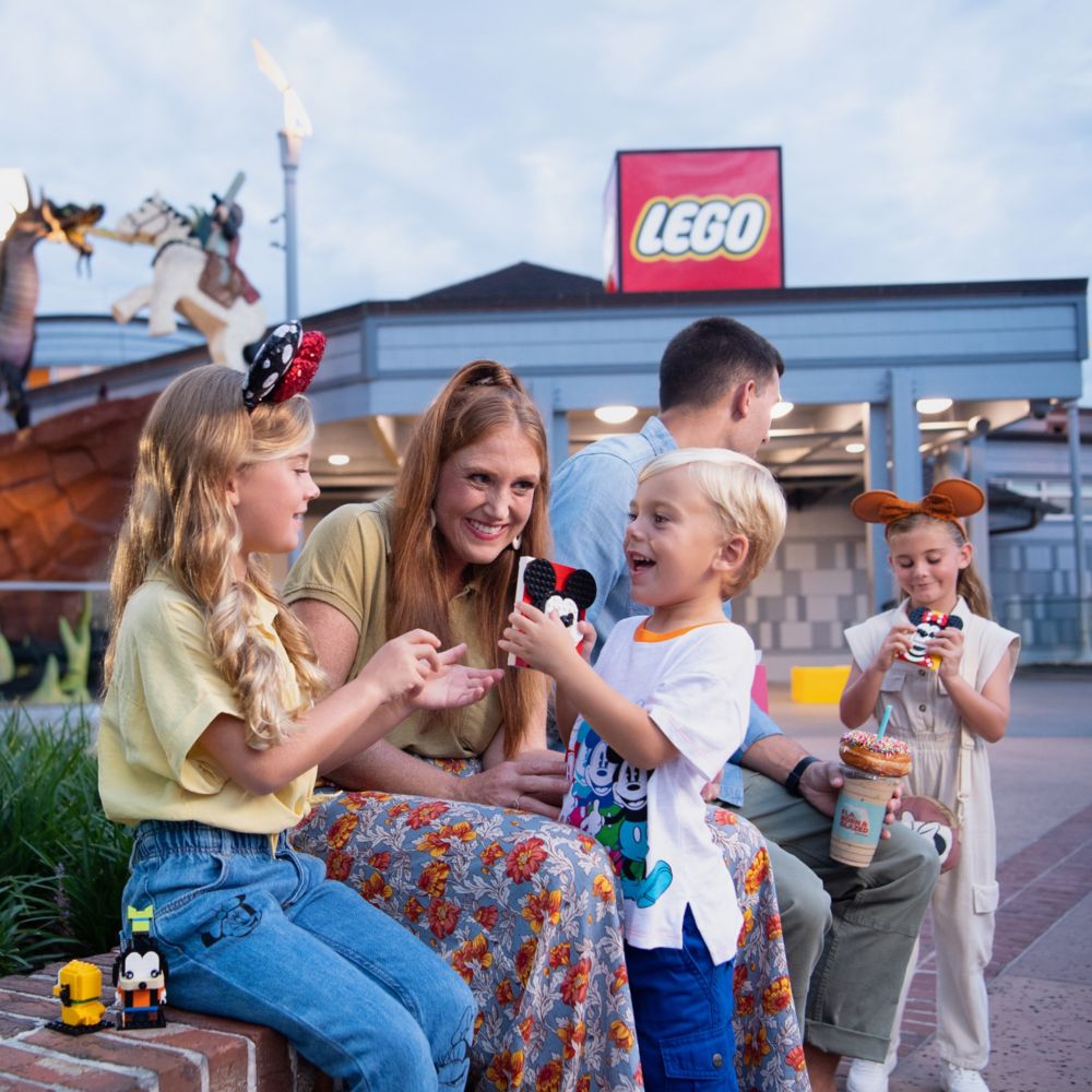 A family of 5 sit along a low wall outside of The Lego Store in Disney Springs