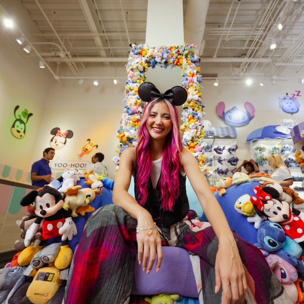 A woman wearing a Minnie Mouse ear headband sitting on a throne decorated with Disney Character plush toys inside the Disney Ever After store