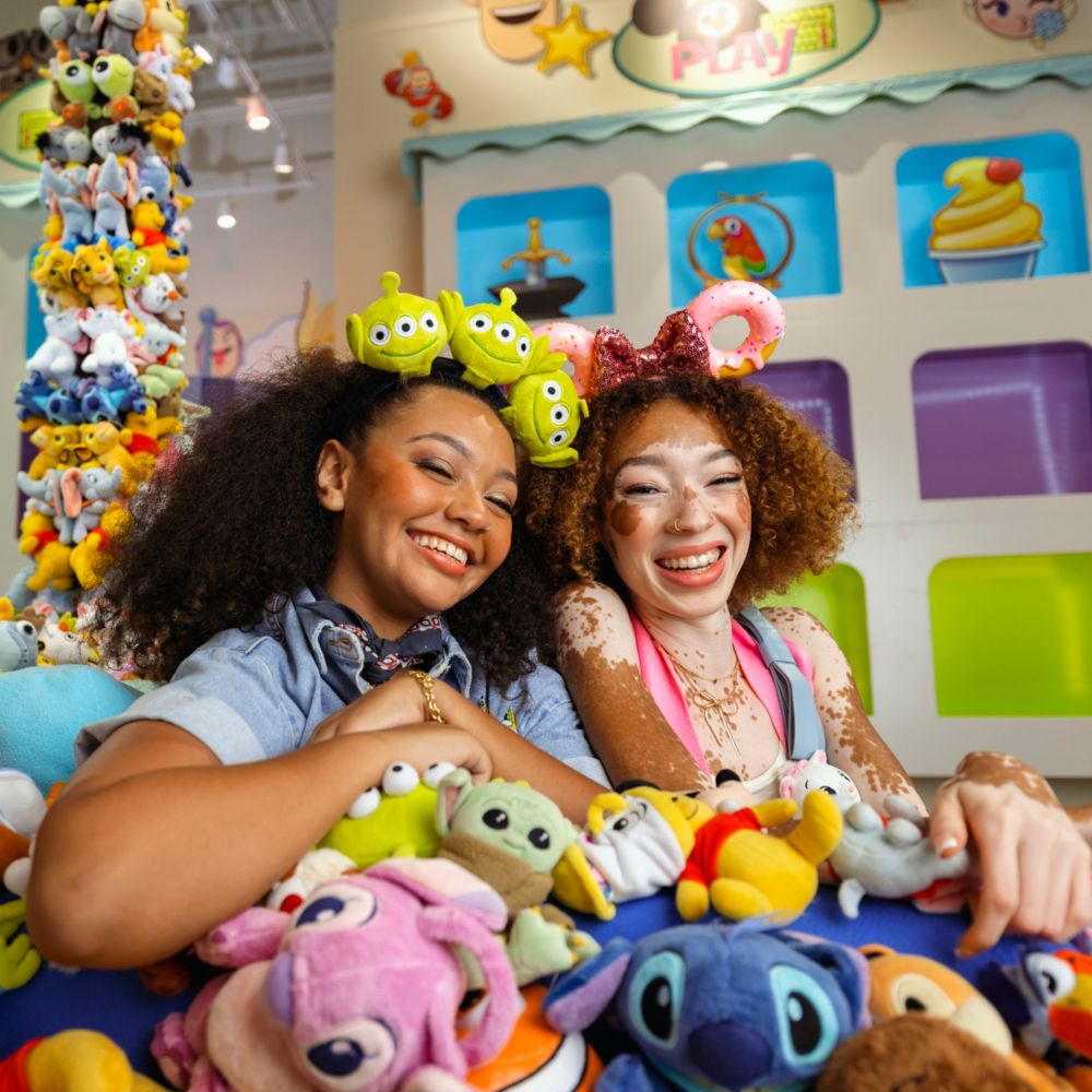 Two women wearing a Toy Story themed headband and a donut themed Minnie Mouse ear headband sitting on a throne of Disney plush toys inside the Disney Ever After store