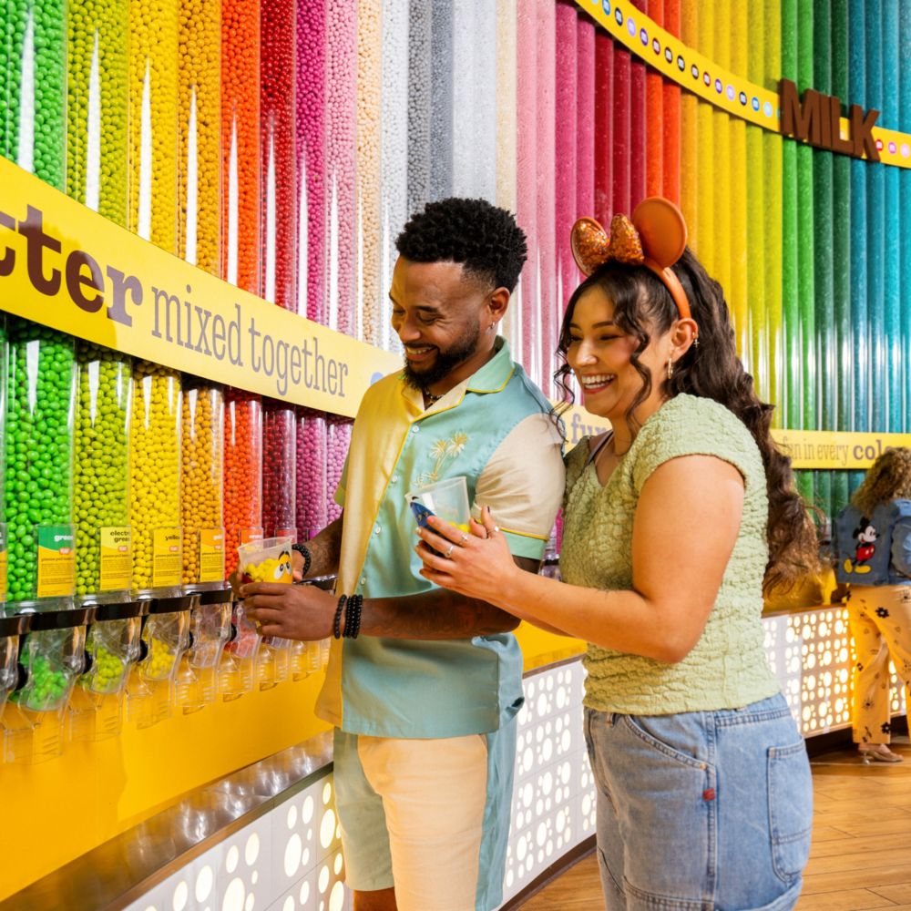 A man and a woman in Disney themed apparel collecting M & M’s from a store with a wall of M & M’s dispensers