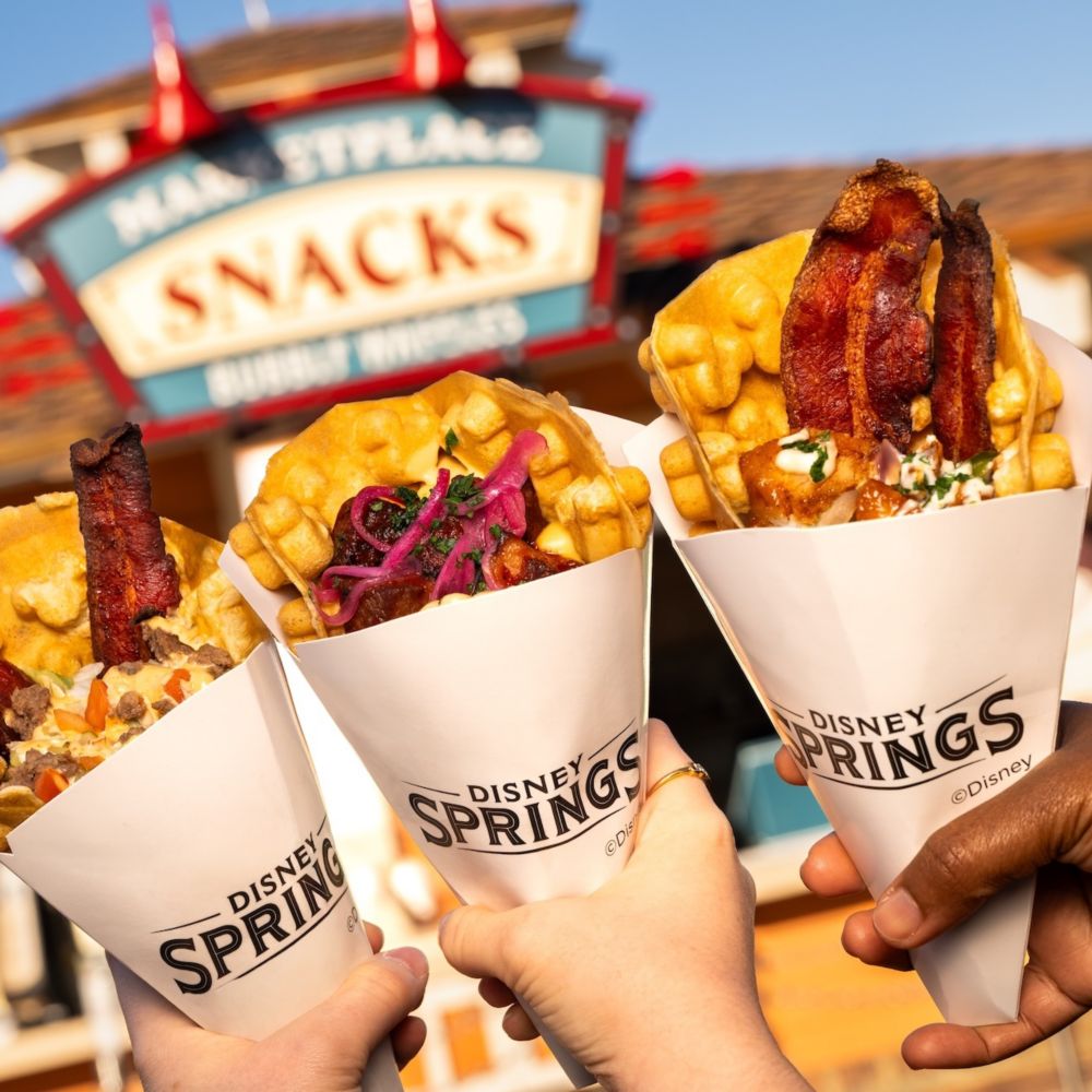 Three hands holding up savory waffle cones in front of Marketplace Snacks at Disney Springs