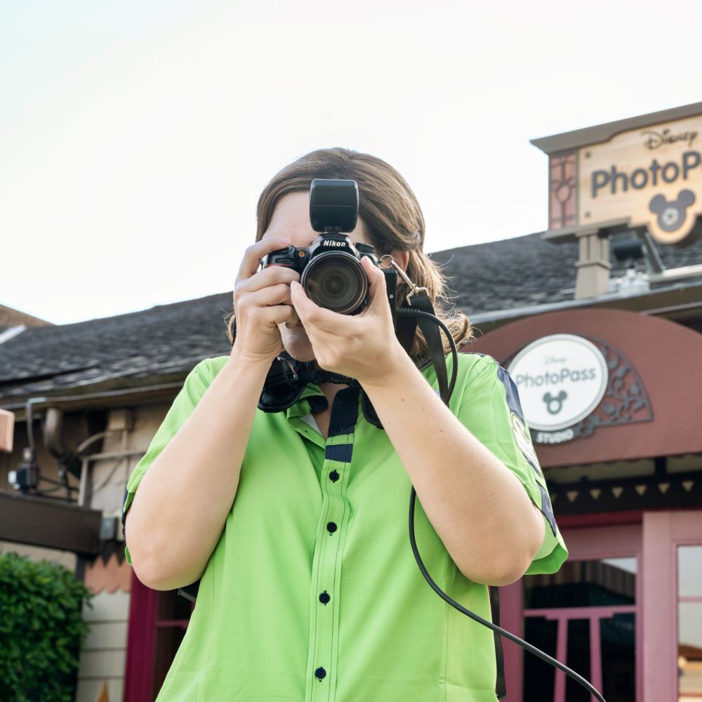 A woman stands outside a building with a sign that reads ‘Disney PhotoPass,’ taking a photo with her camera