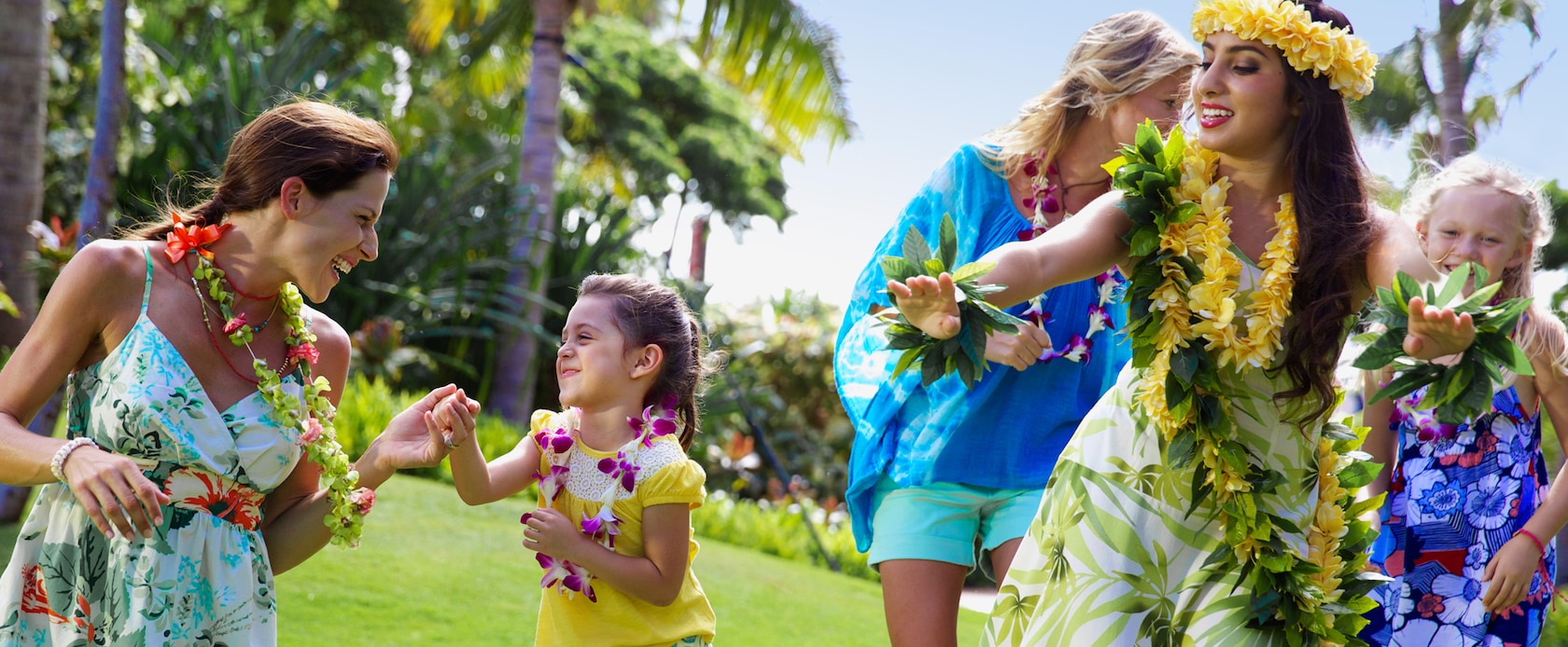 Two young girls and their mothers wearing flower leis follow along during a Hawaiian hula dancing lesson