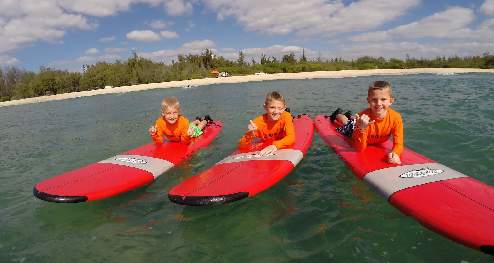 Three young boys give the hang loose hand sign as they lie on their bellies on surfboards floating in the ocean