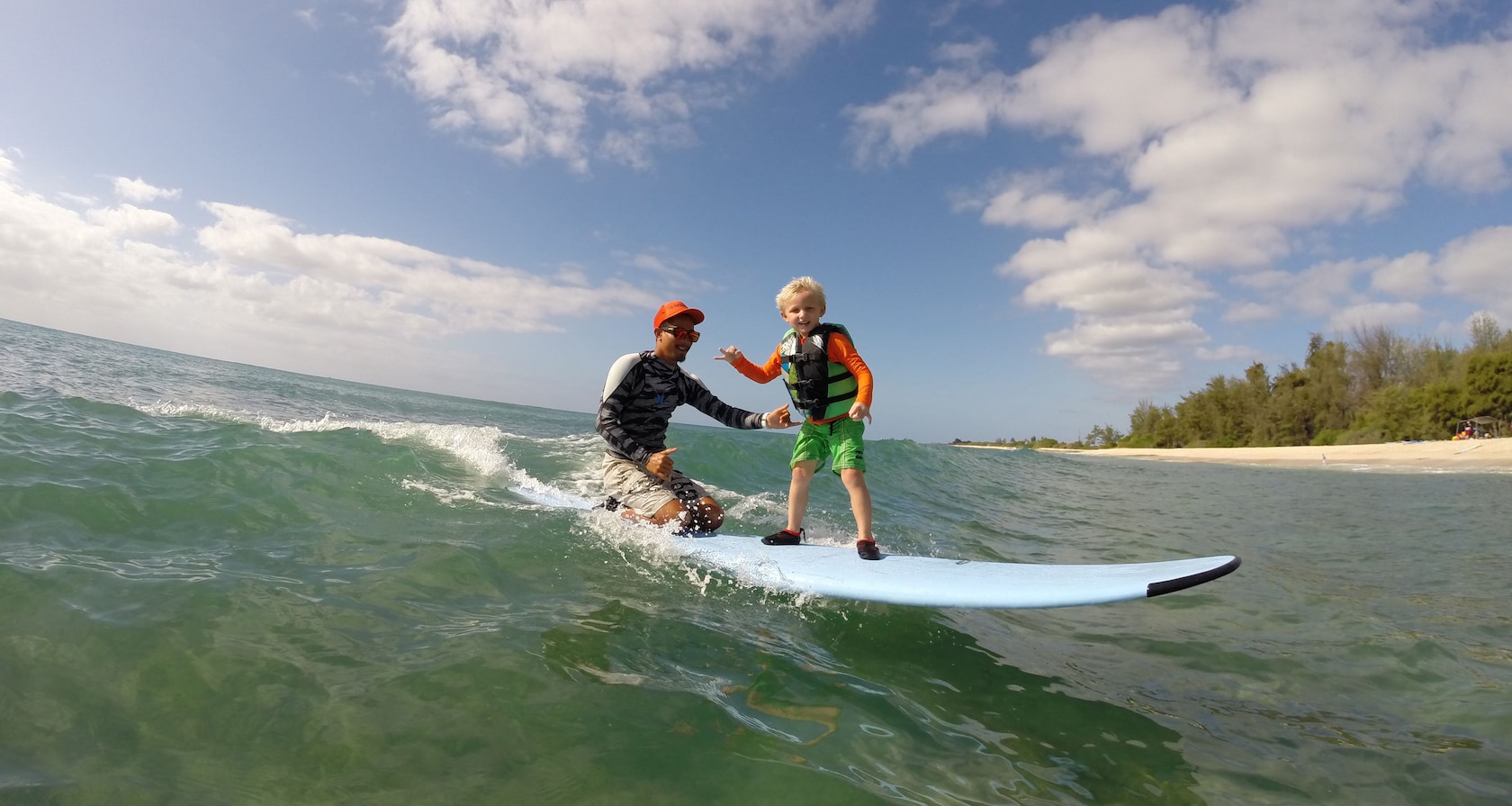 A young boy stands and an instructor kneels on a surfboard atop a breaking wave
