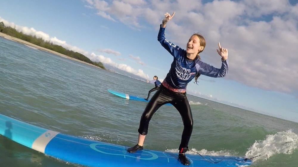 A teenage girl rides a surfboard in the ocean and gestures gleefully