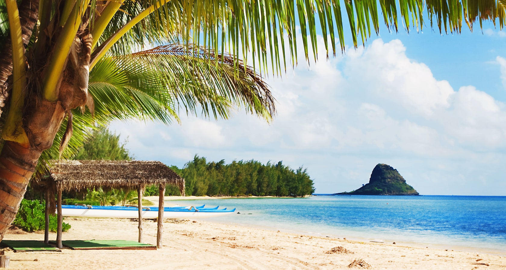 The shoreline of the secret beach overlooking Kāneʻohe Bay and Mokoli`i island with multi person canoes and a beach hut
