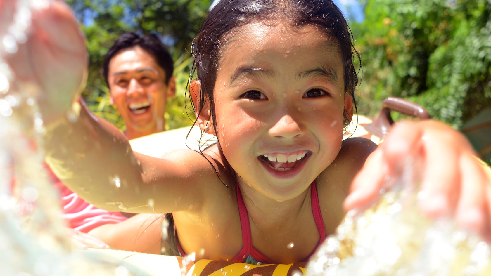 A laughing young girl wet from splashing rides an inflated tube as his father looks on laughing behind her.
