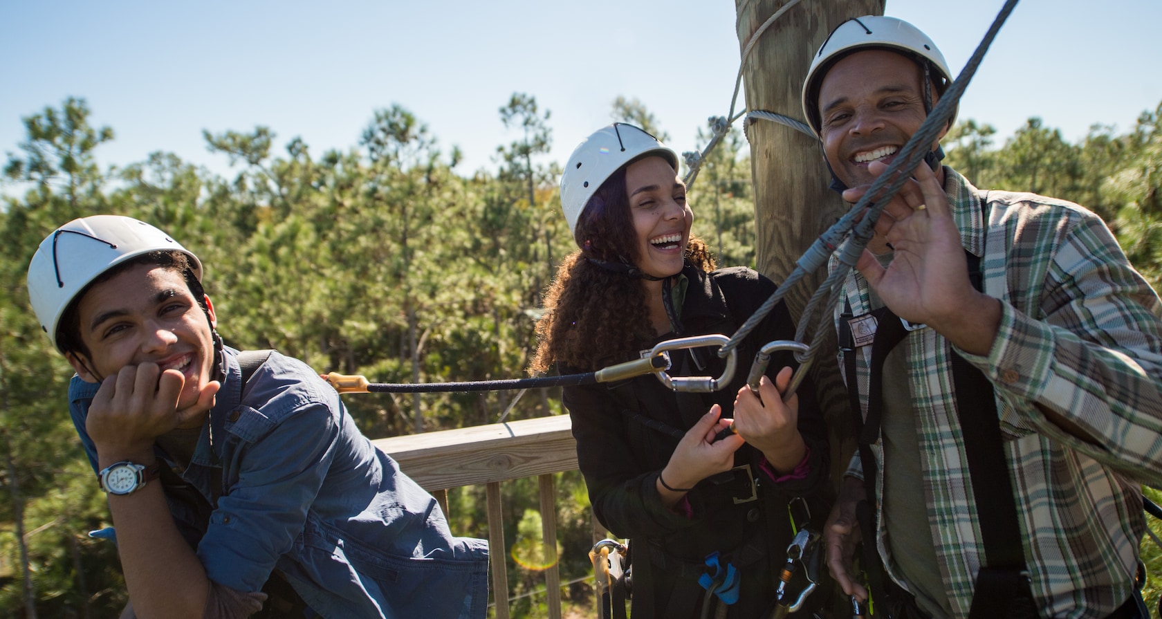 Two young men and a young woman in helmets wear harnesses attached to a zip line