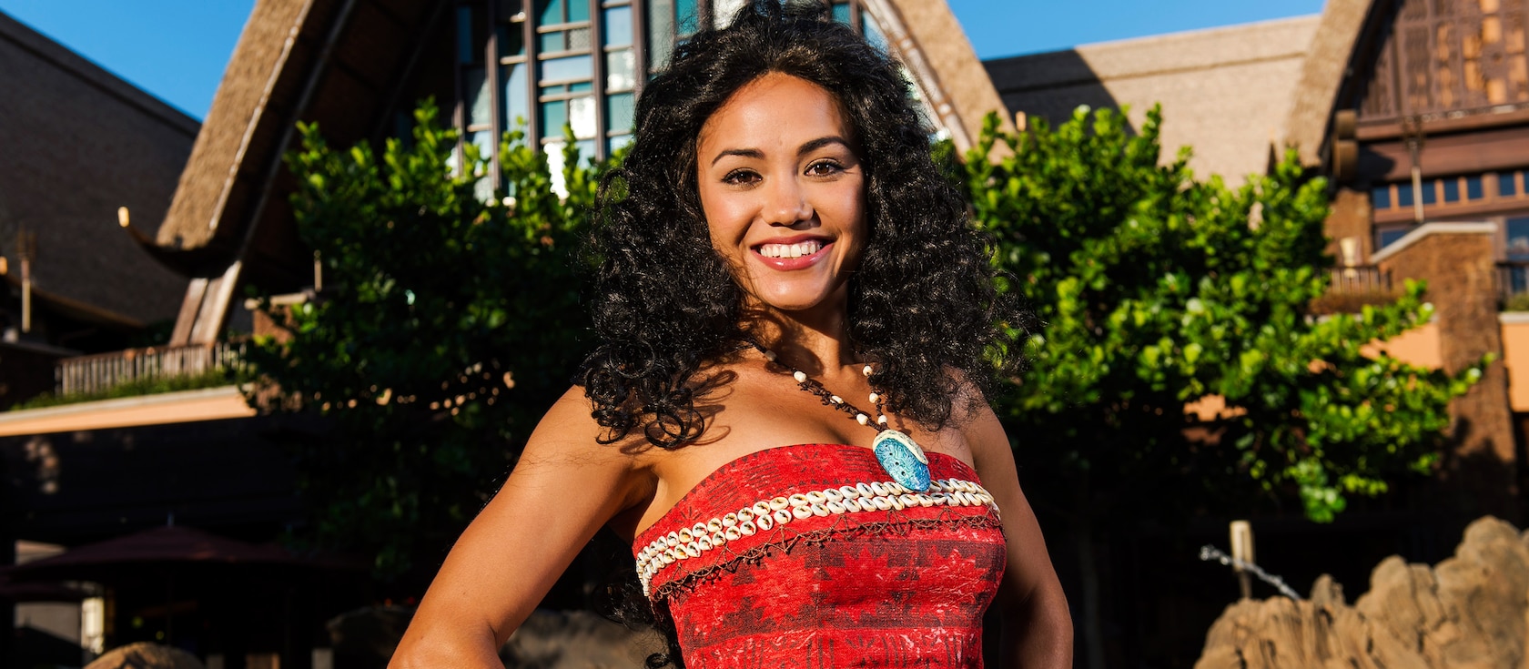 The Moana Character stands smiling in front of Aulani, wearing a top emblazoned with traditional island patterns and small seashells. Her signature necklace, with seashell charm, hangs around her neck.