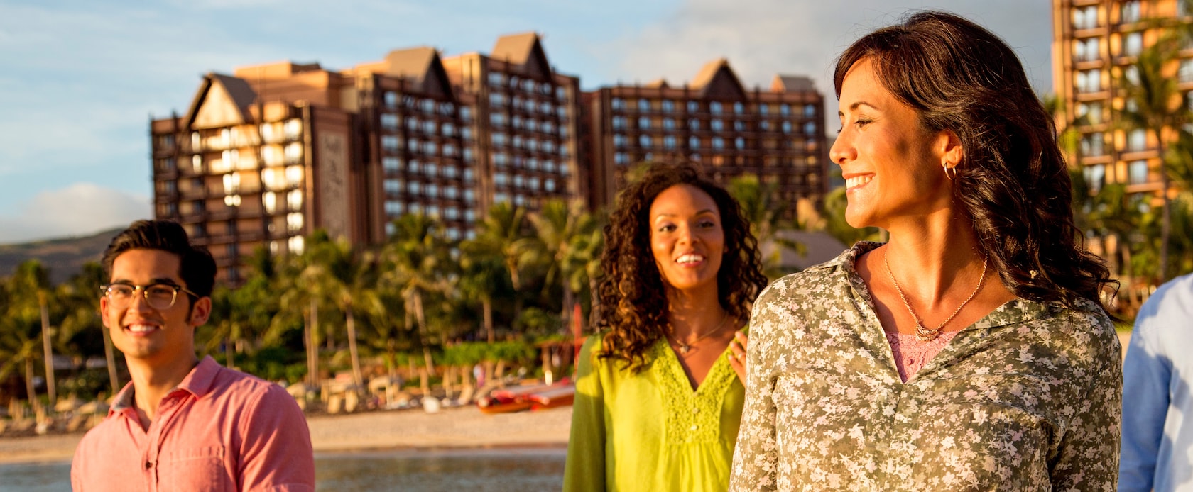 A young man and two young women walk along the beach in front of the Aulani Resort.