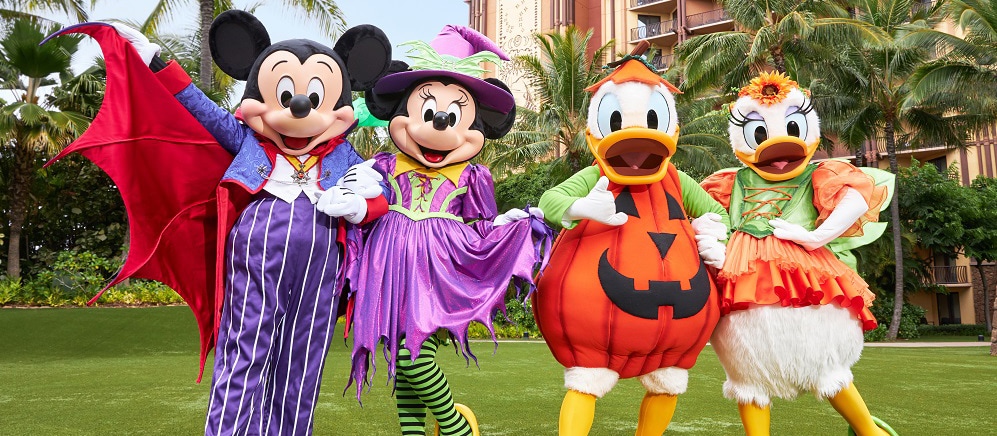Mickey Mouse and Minnie, dressed in purple Halloween costumes, stand next to Donald Duck and Daisy, in pumpkin costumes