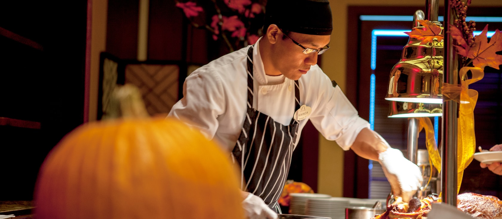 A chef wearing an apron and bandana puts the finishing touches on a dish at a serving station decorated with Thanksgiving décor that includes a pumpkin