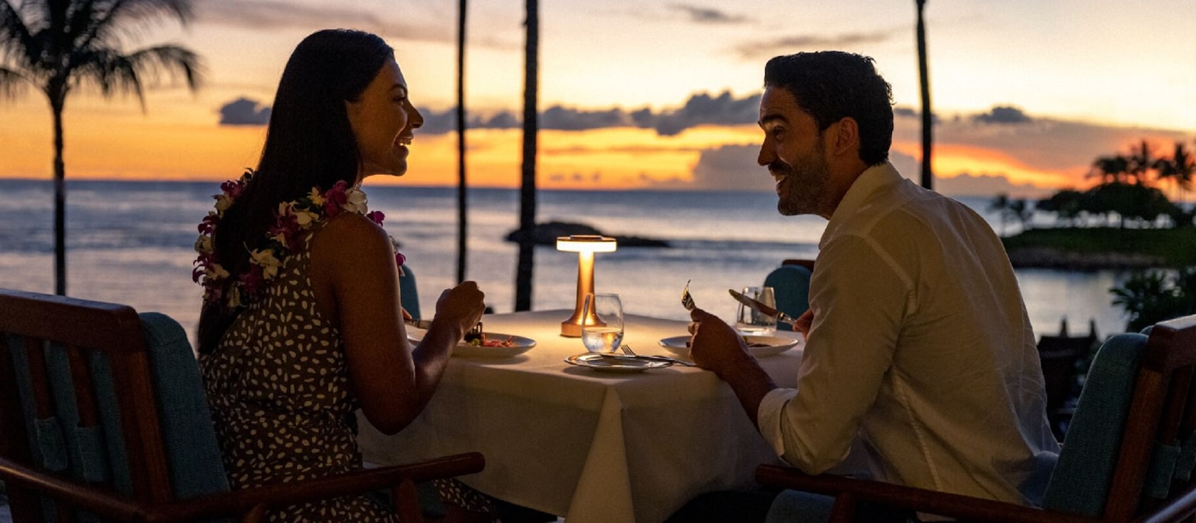 A man and woman sit outside at a table by the ocean, enjoying a romantic dinner at sunset