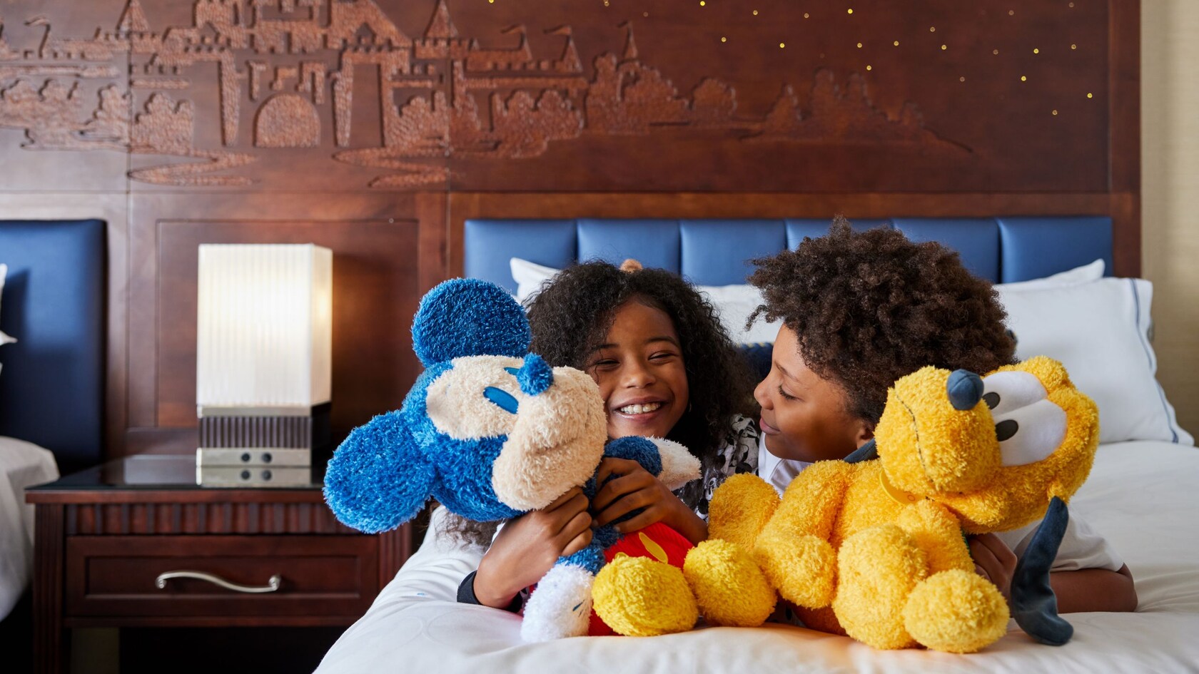 Two children smile and cuddle Mickey Mouse and Pluto plush toys while relaxing on a hotel bed at the Disneyland Hotel