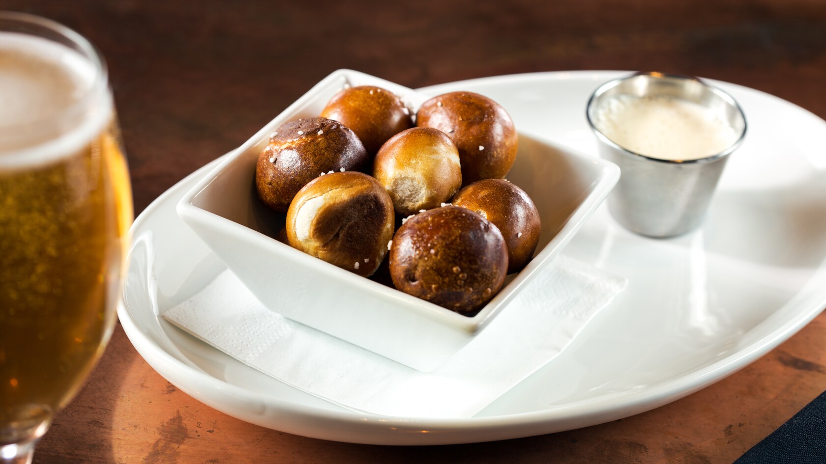 Um copo de chope ao lado de uma porção de petiscos de Pretzel Bites e acompanhamento de molho de queijo gouda com IPA