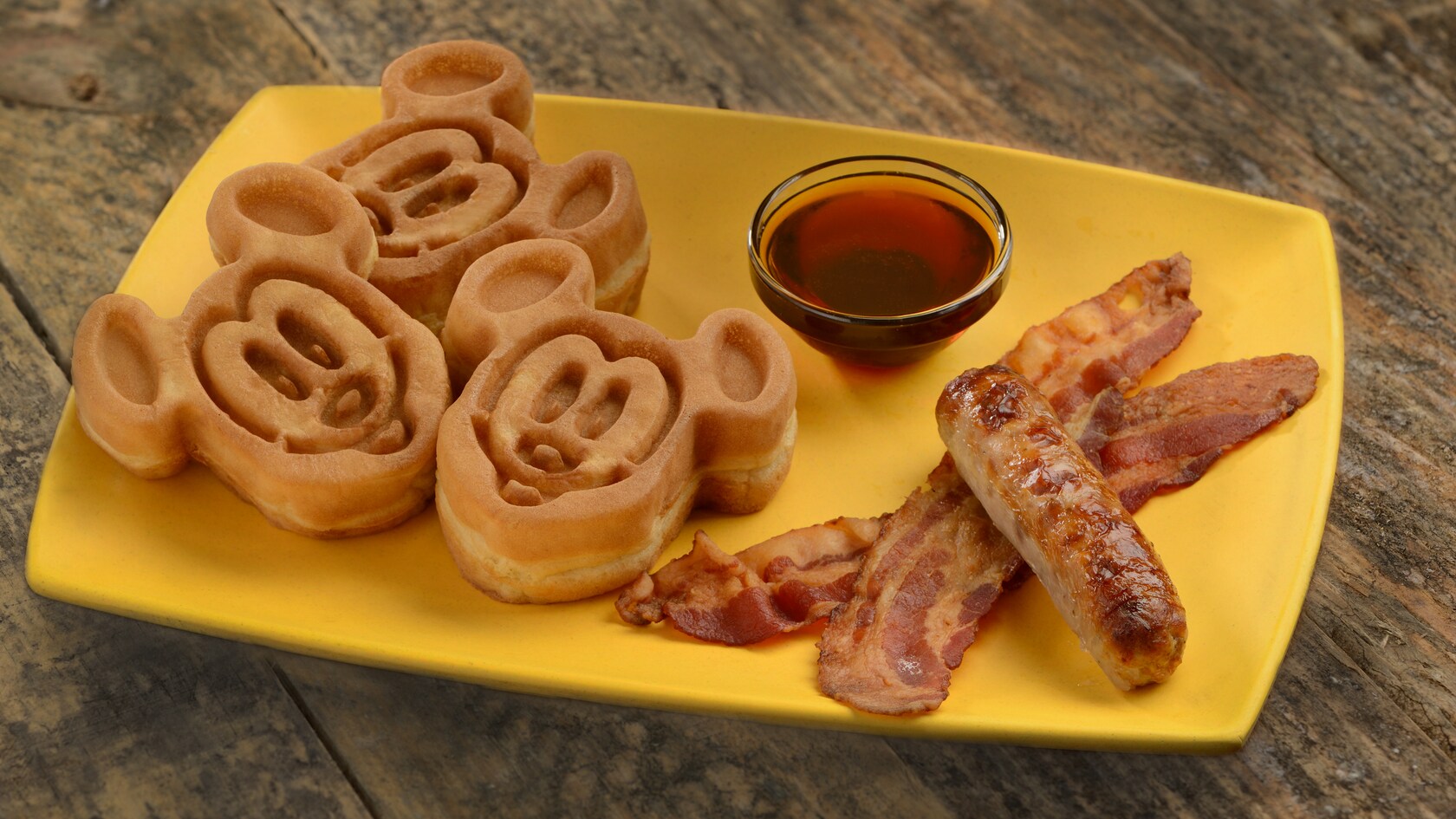 Three waffles in the shape of Mickey Mouse, a bowl of syrup, bacon and sausage on a plate