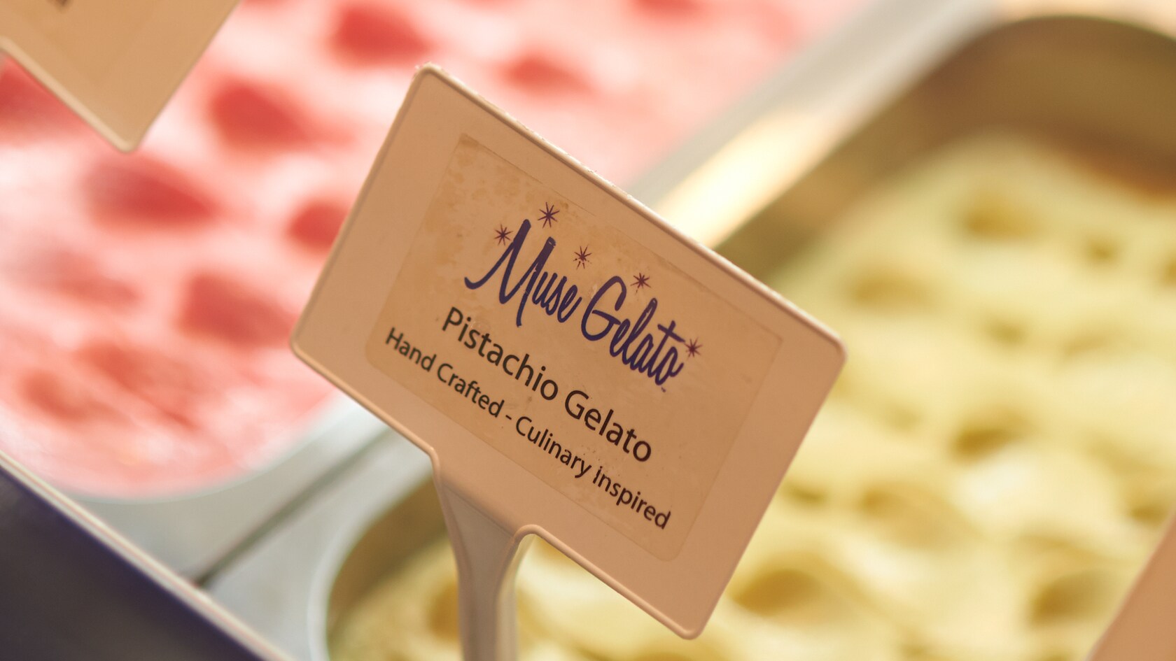 A metal container filled with Gelato in a frozen case and a sign that reads Pistachio Gelato, handcrafted and culinary inspired